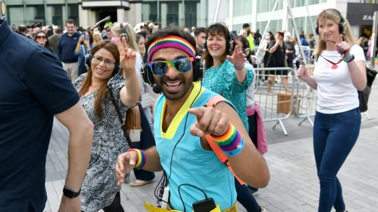 A man is pictured in sunglasses, a blue sleeveless top, a multi-coloured headband, sweatbands and a cassette player. He is walking past Birmingham Bull Ring with crowds of people behind him. 