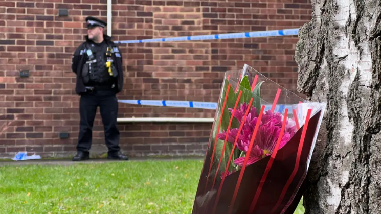 A police officer stands next to a brick wall and overlooks the scene. There are two lines of police tape next to him which is blue and white. In the foreground, a tree trunk has a bunch of purple flowers leaning up against it.