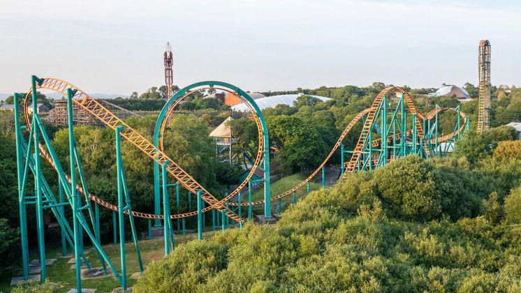 A steel orange and green rollercoaster at Oakwood Theme Park. It is surrounded by green trees and in the background you can see other rides ad buildings.