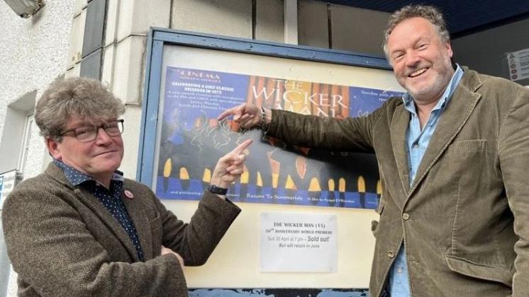 Two men in their 50s either side of a poster for The Wicker Man outside a cinema. Dominic, on the left, is in a tweed suit and has glasses and thick hair. Justin (right) is in a cord suit and has a beard. They are smiling and pointing at the poster.