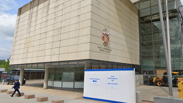 A big beige building is on the left side of the picture, with a glass building adjoined on the right. A Badge with writing underneath says South Cambridgeshire District Council. 