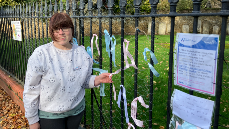 Adele standing next to a fence with pink blue and white ribbons and a note saying Baby Loss Awareness Week