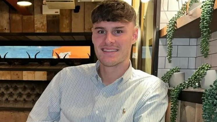 A young man with brown hair, wears a smart shirt and smiles at the camera, sitting in a restaurant 