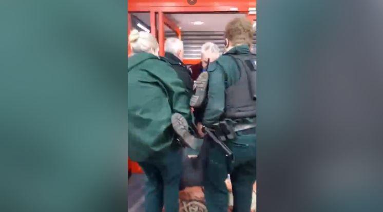 Officers in uniform and a man carry a woman out of a shop. Her legs and feet are being held up by the officers who are carrying her. Shutters are partially down at the doors of the shop.