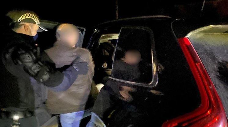 An image of a man, with his face blurred, being ushered into the back of a police car by an officer. It is dark.