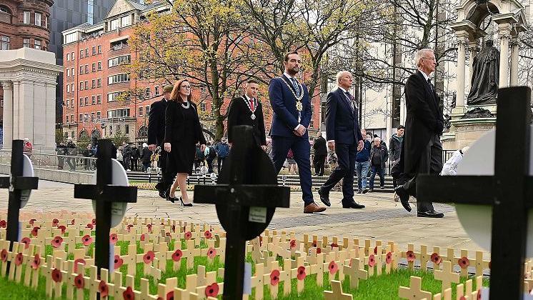 Belfast Lord Mayor Mickey Murray, wearing a navy suit and his chains of office, walks in the grounds of Belfast City Hall with other dignitaries during the Armistice Day event.  Many small wooden crosses with poppies have been left in the grass as a tribute to fallen soldiers.