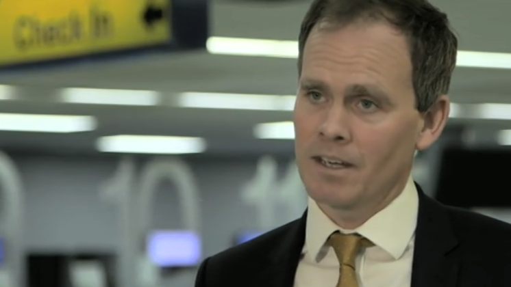 A man with dark hair wearing a dark suit, white shirt and brown tie faces the camera as he is interviewed.