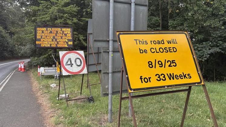 Roadwork signs line the side of a road. One yellow one reads 'This road will be closed 9/9/25 for 33 weeks', one shows a 40mph limit and a digital screen explains a diversion that will be in place. There are some red and white cones to the side of the road.