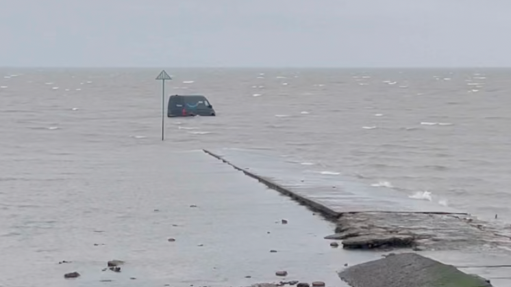 A large, blue Amazon delivery van is partially submerged in thick, brown mud on a wide, flat expanse of coastal terrain.