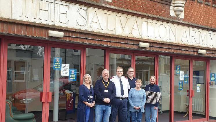 Six people standing outside a red building. The Salvation Army is inscribed into stonework in huge lettering on the front of the building.