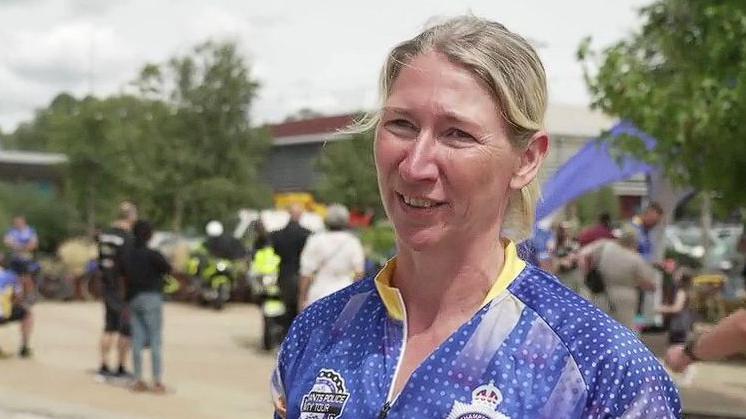 Lorna Clarke with blond hair tied back. She is looking to the left of the camera. She is wearing a blue top with a Northamptonshire Police logo. There are people behind her and what appears to be a blue inflatable slide. There are trees to the left and right.