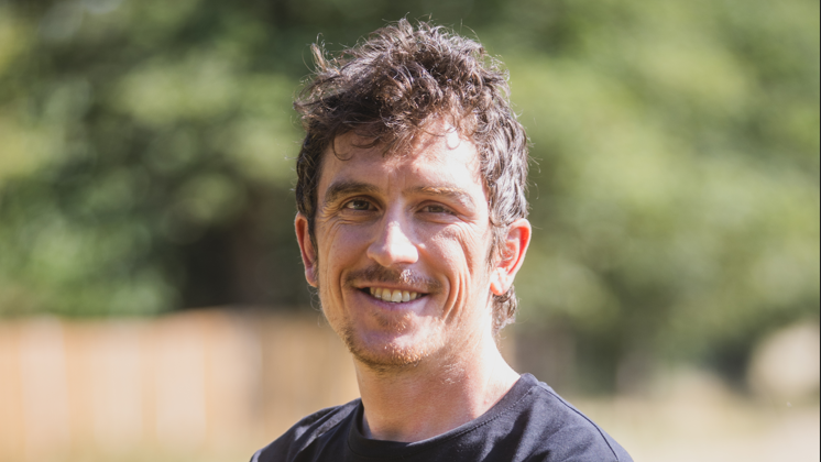 Geraint Thomas is in the middle of the image looking towards the camera smiling. He has short brown hair and a short moustache and beard and is wearing a black T-shirt.