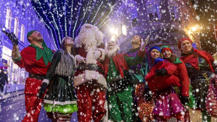 A group of people dressed as Christmas elves and Father Christmas stood in a line in St Peter Port. The Christmas lights are lit up above them in blue light. Fake snow is falling around them. 