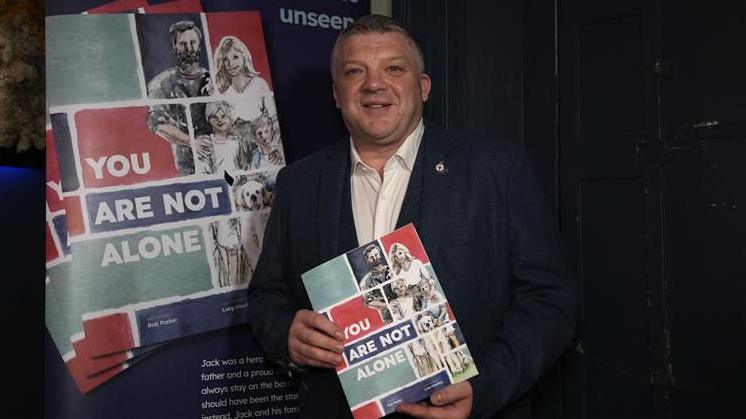Nigel Seaman smiles at the camera as he holds the new book. He has short dark and grey hair and wears a navy suit jacket with a white shirt underneath. A poster board promoting the new book stands behind him.