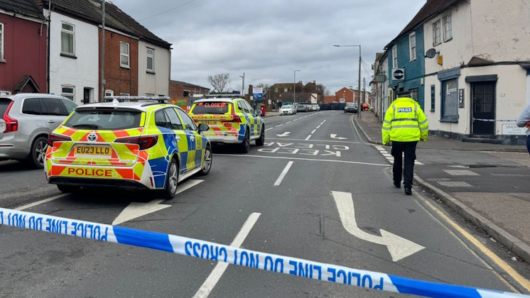 Two Essex Police cars are on the left of a road behind blue and white police tape. A section of the road has been cordoned off. A police officer is walking away from the camera on the right side of the road.