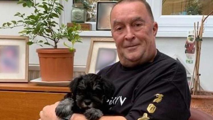 Tony Hughes, is holding a small black puppy and wearing a black jumper. He has very short black hair.