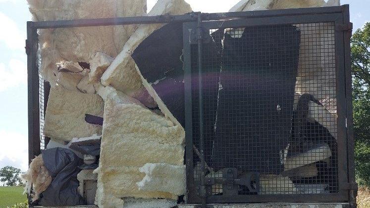 Waste crammed into the back of a lorry trailer, with mesh fencing, showing foam items. Trees and bushes are around the lorry.