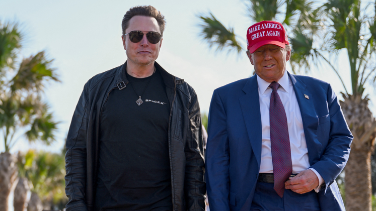 Tesla chief executive Elon Musk wearing a black top and black leather jacket, standing next to US President Donald Trump who is wearing a blue suit, purple tie and red hat with the words make America Great Again in white letters on the hat