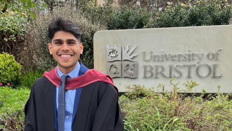 South Asian man smiling at the camera while wearing his graduation robes.