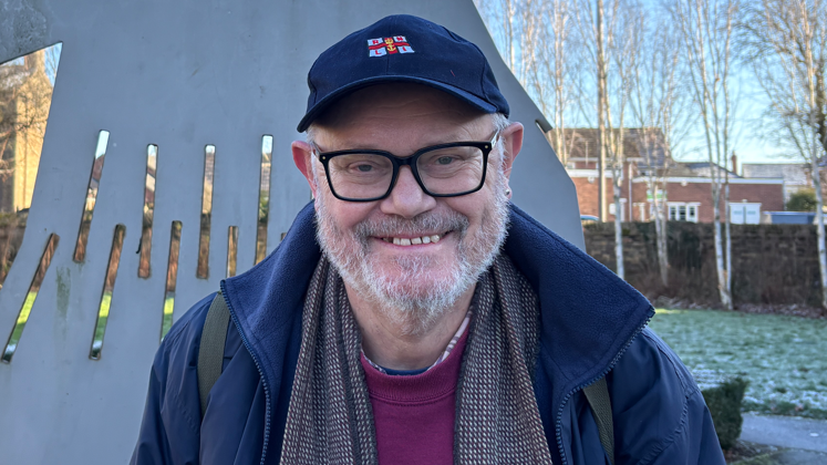 Brian Kell is smiling directly at the camera while standing in front of a metal 2D Straw Bear sculpture. He has black square framed glasses and grey facial hair. He wears a blue cap with the RNLI's lifeboats logo in the centre. He also wears a blue coat and red jumper - both branded with a Straw Bear.