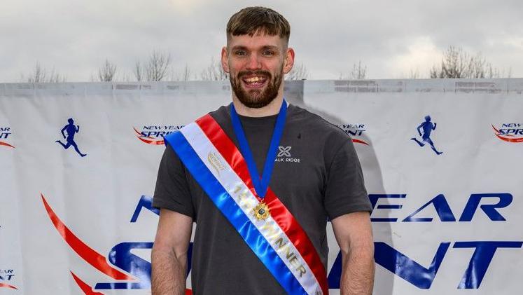 Happy athlete with short brown hair and short beard in grey T-shirt sports red, white and blue race victor sash across chest and  winner's gold medal.