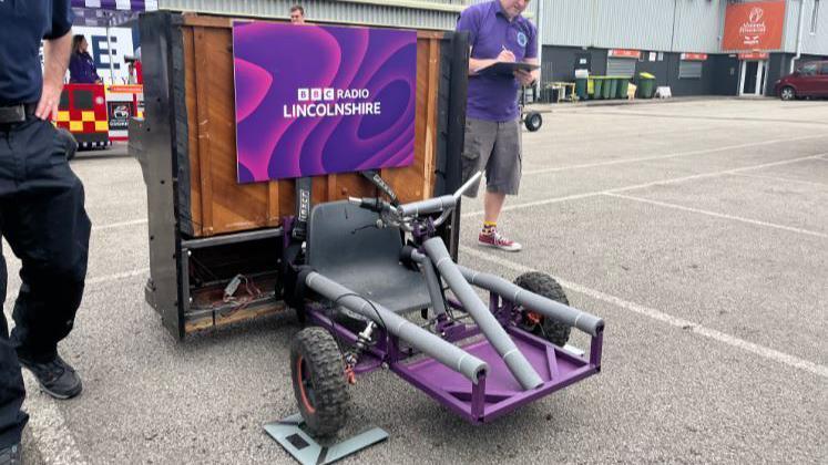 BBC Radio Lincolnshire soapbox with a piano strapped to the back with a purple BBC Radio Lincolnshire sign and a seat with two front wheels