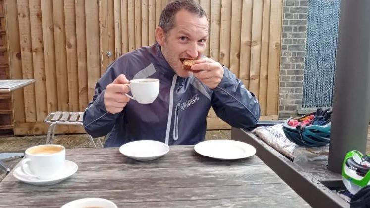 A man sitting at a wooden table wearing a blue jacket. He has short dark hair. He is eating a cake with his left hand and holding a coffee cup in his right hand. There are other cups and plates on the table. Next to the table are cycling helmets.