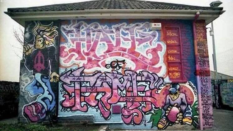An early graffiti piece called ' Hall of Fame' by INKIE and FELIX painted onto the side of one of the Barton Hill Youth Club buildings. The piece is very intricate with barely legible words done in pink and purple hues.