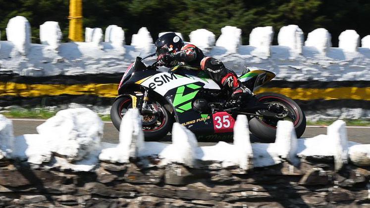 Garry Broughton riding a black motorcycle which has green and white branding on it and the number 35 on a red background. There are white painted stone walls on either side of the course.