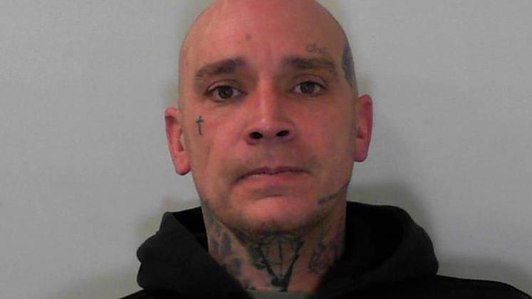 A bald man with tattoos on his face and neck looks at the camera. He is wearing a grey/green t-shirt under a dark zipped-up hoodie.