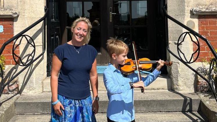 Annabel Thwaite with her son outside Framlingham College. She has blonde hair and is wearing a blue T-shirt and skirt and is smiling. Her son is playing the violin