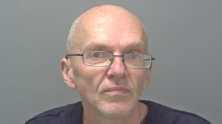 A mugshot of Roy Barclay - a bald man wearing glasses. He is standing against a grey backdrop and is looking directly into the camera.