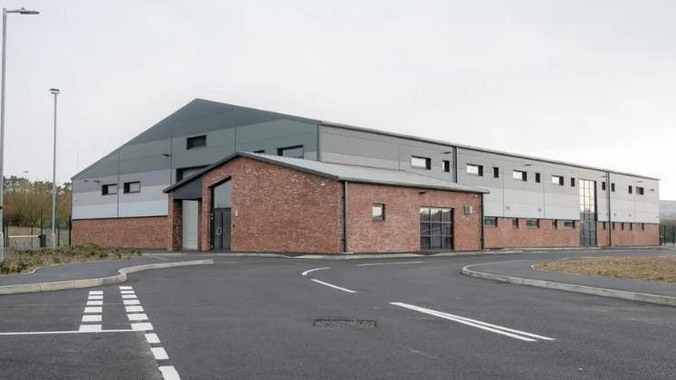 A large building is a mix of grey panelling and red bricks, with a road winding in front of it to the right.