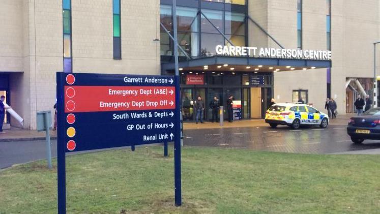 The entrance to the emergency department at Ipswich Hospital. A sign saying "Garrett Anderson Centre" is over the main door. There are people milling about and a police car. In the foreground there is a hospital sign with directions to wards and departments. 