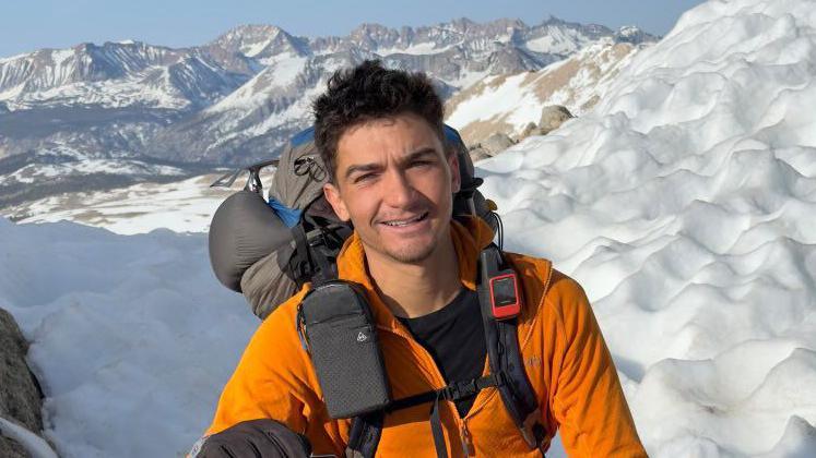 A man with short dark spiky hair looks into the camera. He wears an orange jacket with the top of a grey backpack poking out behind his head. Behind him are snow-covered mountains