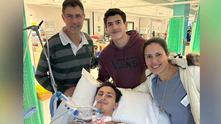 Seb on a hospital bed has a hospital gown on, short black hair and a tube on his nose and throat. His mother is on his left, brother is above him, and his father on his left. The image has been taken on a hospital ward.