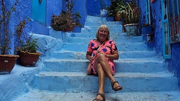 Lynn Stephenson, wearing a flowery dress and sandals, sitting outside on some blue-coloured steps