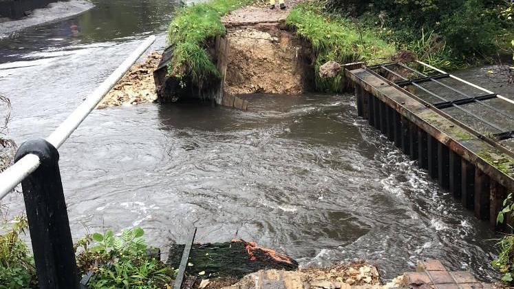 A photo taken from one side of a small weir looking towards a footpath on the other side of the bank