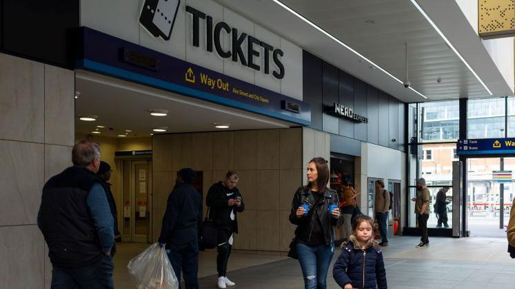 Group of people, some carrying takeaway coffee cups, walking in front of a sign stating tickets in black lettering on a white background