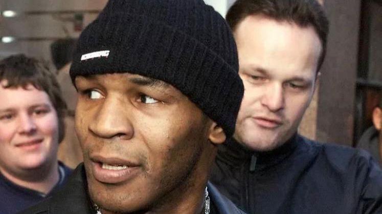 Mike Tyson in a beanie hat flanked by a fan and a security guard in a black jacket 