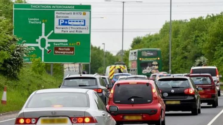 Two lanes of vehicles approach the roundabout, with a large sign for the 'Thickthorn Interchange' to the left. Trees and grass line each side of the road.