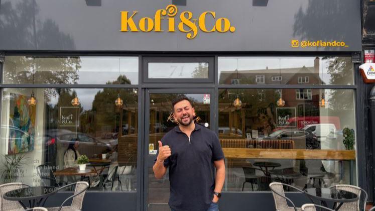 Harry Limbert stands doing a thumbs up gesture in front of a cafe called Kofi & Co