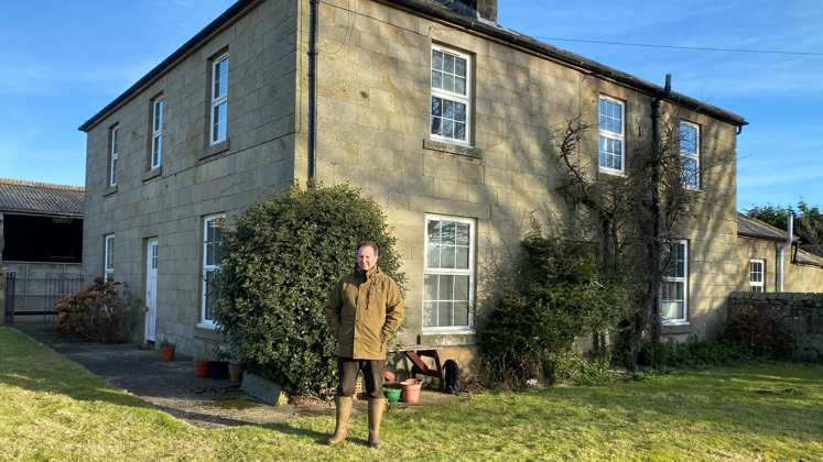 Louis Fell, wearing wellington boots and a green jacket, stands in front of a large Georgian farmhouse. It is rectangular with large windows and a chimney and has bushes around the door. 