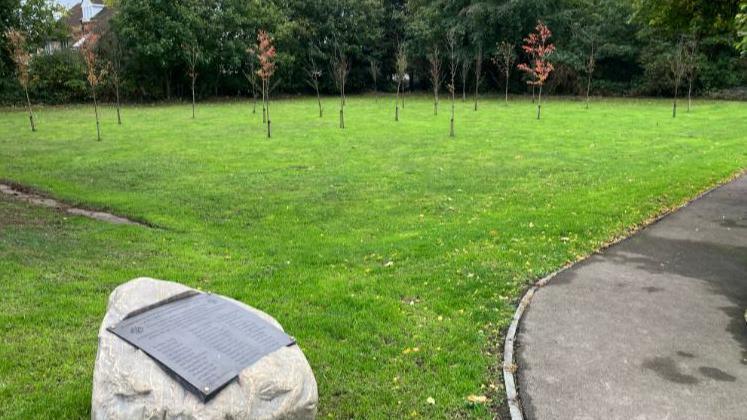 A stone and brass tablet next to a path in a lawned area that is surrounded by large trees. In the centre are two rows of tree saplings.