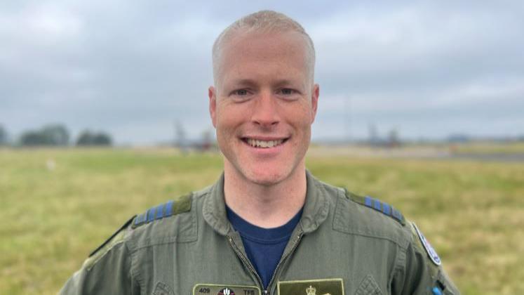 Lieutenant Colonel Ben Switzer smiles into the camera. He has short blond hair and is wearing a green RCAF uniform. He is standing on a green field with two planes in the background