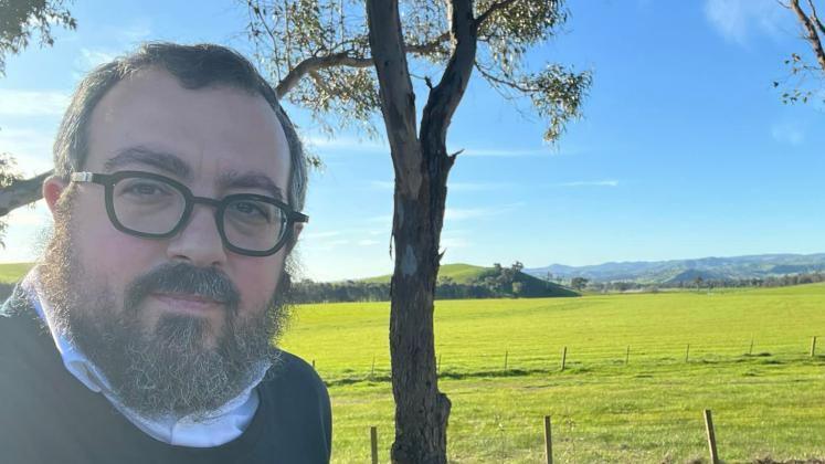 A middle aged man with glasses looks at the camera, behind him is a grassy field.