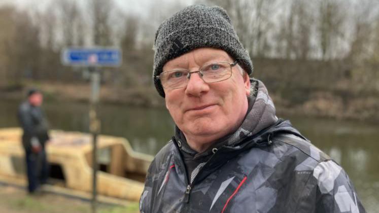 Ian Platt stands by the River Aire, with a small boat in the background. He is wearing a woolly hat, waterproof jacket and glasses.