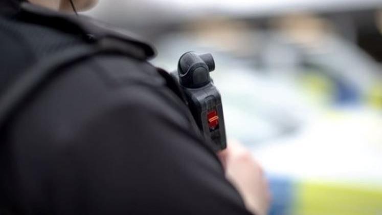 A close up of a police officer's shoulder. They are wearing a bullet proof vest with a radio mounted on it. A police car is blurred in the background.