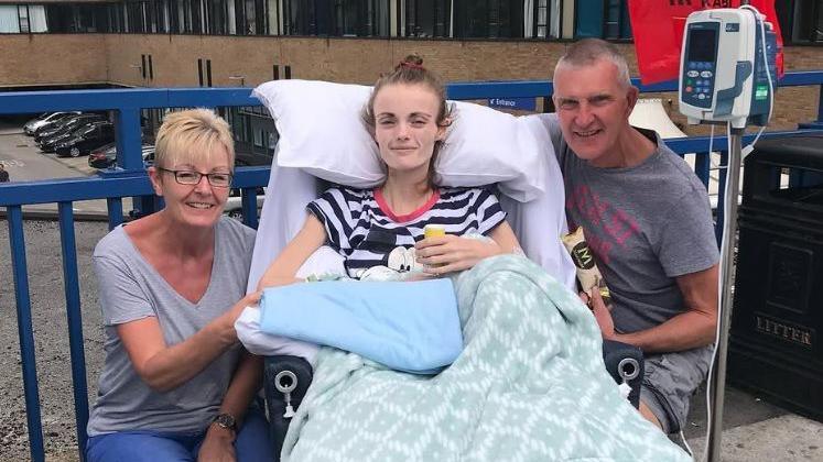 A frail-looking young woman is sitting up in a hospital bed, which has been wheeled on to a balcony along with a portable hospital monitor. She wears a striped T-shirt and is partially covered by a green patterned blanket. Her parents, a man and a woman, are crouched down to either side of her and smiling.