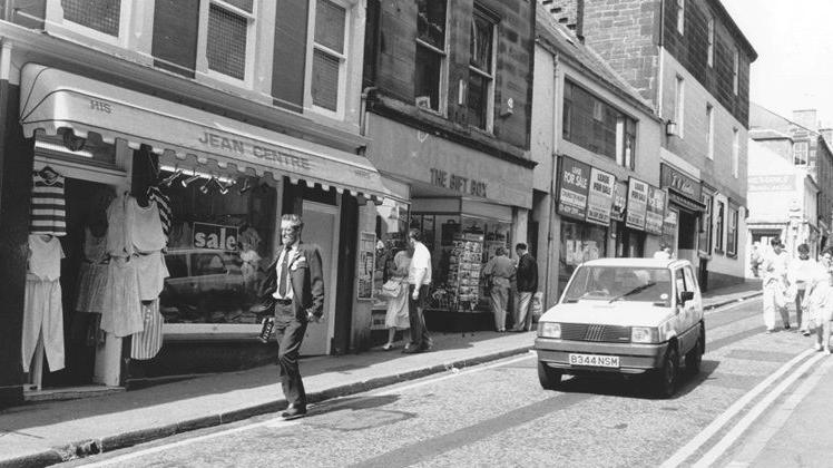 An old image of Friars Vennel in black and white with people walking up the street and a Fiat Panda coming down the street